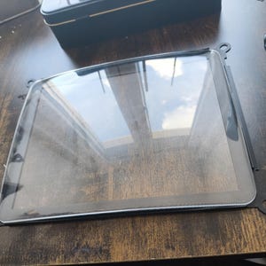 以下が含まれることがあります： 黒いフレームと丸みを帯びた角を持つ長方形の透明なガラススクリーン。スクリーンは空と雲を反射しています。フレームには、各コーナーに小さな円形の取り付けポイントがあります。スクリーンは濃い木材の表面にあります。