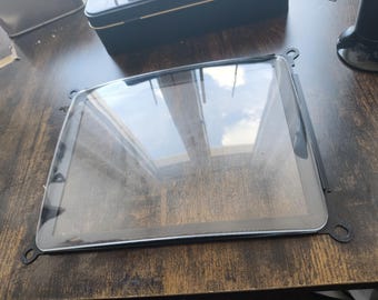 Może przedstawiać: Prostokątny, przezroczysty szklany ekran z czarną ramką i zaokrąglonymi rogami. Ekran odbija niebo i chmury. Rama ma małe, okrągłe punkty mocowania na każdym rogu. Ekran leży na ciemnej drewnianej powierzchni.