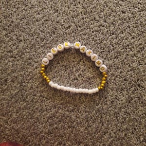 May include: White beaded bracelet with gold letters spelling the name "LEGEMAXIMO".