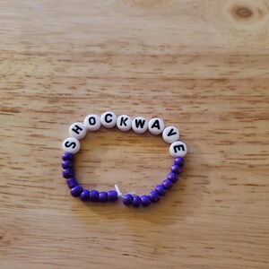 May include: Purple and white beaded bracelet with the word "SHOCKWAVE" spelled out in black letters on the white beads.