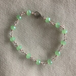 May include: A delicate bracelet featuring light green, faceted beads strung on copper-colored wire. The bracelet is secured with a small clasp. The beads are evenly spaced, creating a simple yet elegant design.