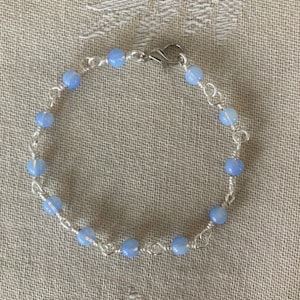 May include: A delicate bracelet featuring light blue beads strung on a silver wire. The bracelet is secured with a silver clasp. The beads are evenly spaced, creating a simple yet elegant design. The jewelry is suitable for everyday wear.