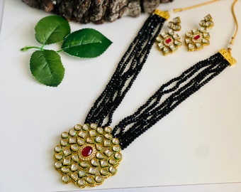 May include: A black beaded necklace with a large, round pendant featuring a red stone surrounded by white and green stones. The necklace is layered with multiple strands of beads and comes with matching earrings.