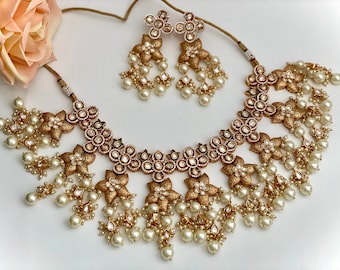 May include: A gold and white necklace and earrings set with pearl accents. The necklace features a floral design with a row of round stones in the center. The earrings are dangle style with a floral design and pearl drops.