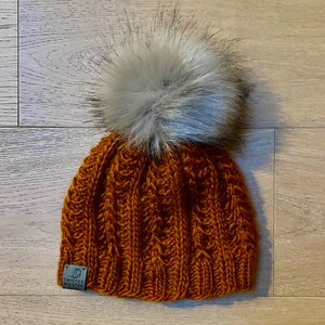 May include: A brown knitted beanie with a white faux fur pom pom. The beanie has a cable knit pattern and a small brown leather tag with the text "Twisted Wisters" on it.