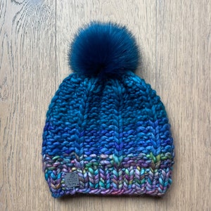 May include: A blue and purple knitted beanie with a fluffy blue pom pom. The beanie has a label that says "Twisted Sister by Chery".