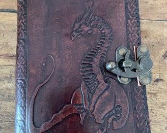 May include: A brown leather journal with a dragon embossed on the front cover. The journal has a metal clasp and a Celtic knot border.