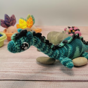 Peut inclure: Un dinosaure crocheté bleu sarcelle et bleu avec des détails noirs, posé sur de petites pierres. La créature faite à la main a de grands yeux expressifs et un aspect texturé. D'autres articles crochetés sont en arrière-plan.