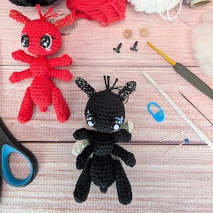May include: Two crocheted amigurumi toys, a red ant and a black ant, with large eyes. The toys are on a wooden surface with craft supplies, including yarn, needles, and a crochet hook.