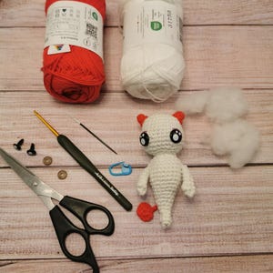 Peut inclure: Scène de bricolage avec fournitures de crochet. Comprend des pelotes de fil rouge et blanc, un crochet, des ciseaux, une petite figurine crochetée avec des accents rouges, des yeux de sécurité noirs et de la ouate. Les articles sont disposés sur une surface en bois.