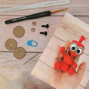 Könnte beinhalten: Ein rotes Häkelroboter-Spielzeug mit grauen Augen und orangefarbenen Akzenten. Das Spielzeug wird in einer Hand gehalten. Das Bild zeigt auch Bastelbedarf wie Garn, Pfeifenreiniger, Sicherheitsaugen und eine Häkelnadel.