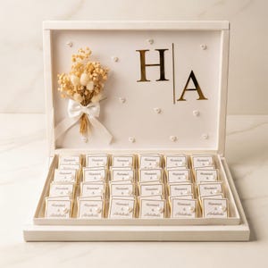 Könnte beinhalten: Eine weiße Schachtel voller Pralinen, verziert mit goldenen Buchstaben und einem kleinen Strauß getrockneter Blumen. Die Schachtel ist mit den goldenen Buchstaben "H" und "A" sowie kleinen Perlenakzenten verziert. Jedes Pralinenquadrat hat Text und eine Perle.