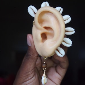 May include: A tan ear model decorated with white seashells and a gold wire earring. A gold wire with brown beads and a white shell pendant hangs from the ear. The ear is held by a hand against a dark background.