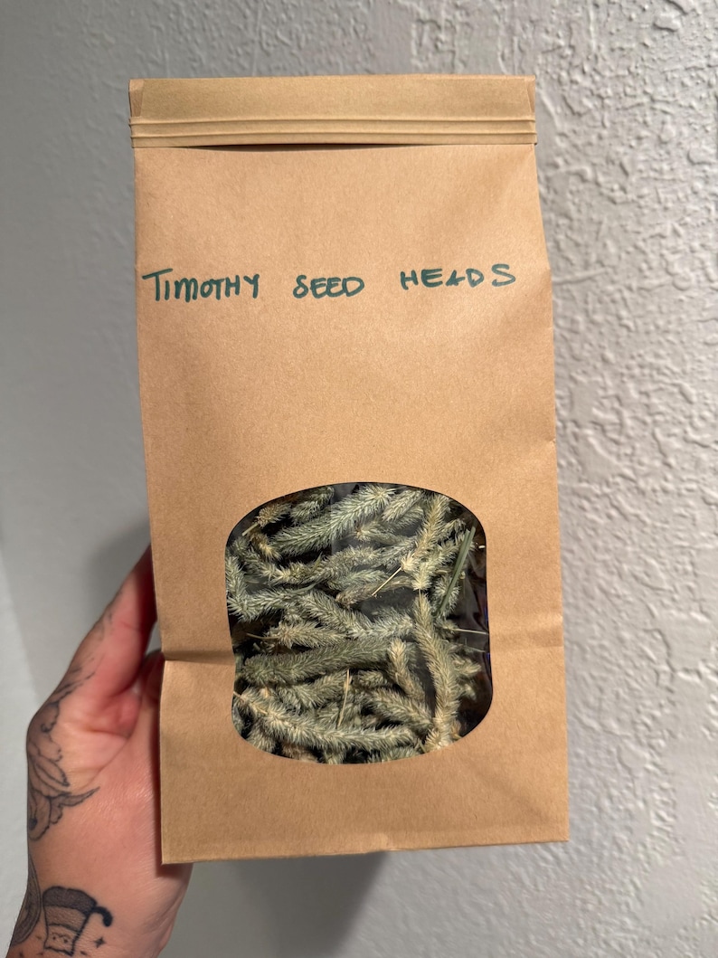 May include: A brown paper bag with a clear window displaying dried Timothy seed heads. The bag is labeled in green marker with the words "Timothy SEED HEADS". The seed heads are a light green color.