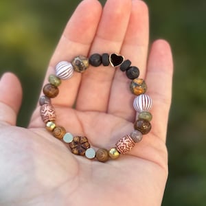 May include: A beaded bracelet featuring a variety of natural stone beads in shades of brown, gold, and green. The bracelet includes striped, patterned, and spherical beads, with a black heart-shaped accent. The bracelet is held in a hand.