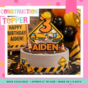 Peut inclure: Un gâteau d'anniversaire sur le thème de la construction, avec une base de glaçage grise, décoré d'une bande d'avertissement jaune et noire. Le gâteau est surmonté d'un décor sur le thème de la construction avec le chiffre 3 et le nom "AIDEN".