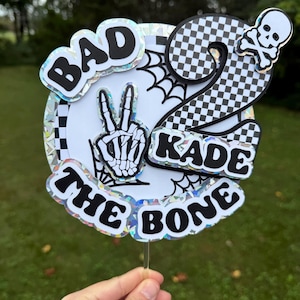 May include: A birthday cake topper with the words "BAD THE BONE" and "KADE" in black, outlined in white. The number "2" is in a checkered pattern, and a skeleton hand makes a peace sign. A skull and crossbones are also present.