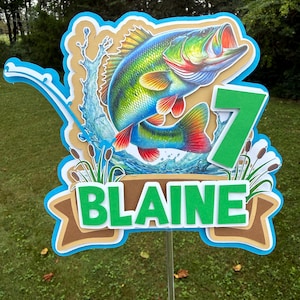 Peut inclure: Décoration de gâteau multicouche avec une illustration colorée d'un achigan à grande bouche sautant hors de l'eau. Le design comprend le chiffre 7 en vert, le nom "BLAINE" en vert et une canne à pêche. La décoration est tenue par un bâton transparent.