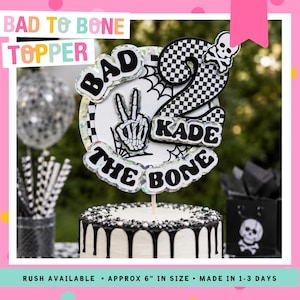 Pode incluir: Um topo de bolo com o texto "BAD TO BONE TOPPER" e "KADE THE BONE". O topo apresenta uma mão de esqueleto fazendo o sinal de paz, um número 2 quadriculado e uma caveira. O bolo é branco com gotas e granulado preto.
