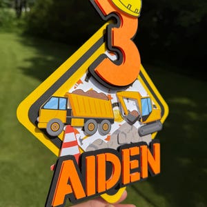 May include: A vibrant birthday cake topper with a construction theme. It features a yellow hard hat, the number 3, a dump truck, an excavator, and the name "AIDEN" in orange and black. The topper is held by a clear stick.