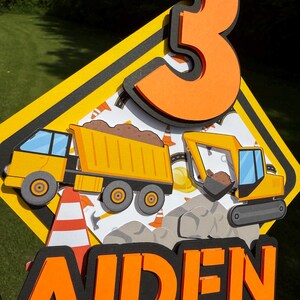 May include: A layered construction-themed cake topper. The design features a yellow and black road sign shape with a large orange number 3, a dump truck, an excavator, and the name "AIDEN" in orange letters.