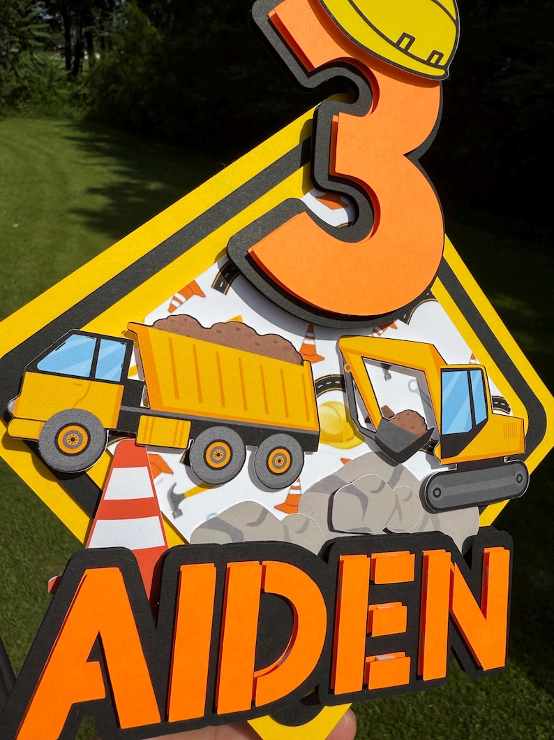 May include: A layered construction-themed cake topper. The design features a yellow and black road sign shape with a large orange number 3, a dump truck, an excavator, and the name "AIDEN" in orange letters.