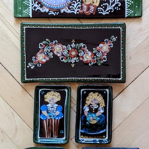 Op de afbeelding: Set van zes handgeschilderde keramische tegels met volkskunstontwerpen. De tegels zijn versierd met bloemmotieven, figuren in traditionele kleding en een eend. De tegels zijn in tinten bruin, groen, blauw, geel en wit.