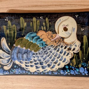 May include: A ceramic dish with a hand-painted design of a white duck with blue and green feathers. The duck is surrounded by green leaves and blue berries on a black background.