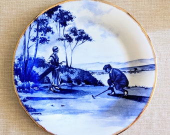 May include: A decorative plate with a blue and white golf scene. The plate features two figures on a golf course, with trees and a body of water in the background. The rim is trimmed with gold.