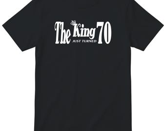 Może przedstawiać: Czarny t-shirt z krótkim rękawem z białym napisem The King JUST TURNED 70 oraz małą grafiką korony umieszczoną nad słowem King. Wzór jest wyśrodkowany na klatce piersiowej koszulki, która posiada okrągły dekolt.