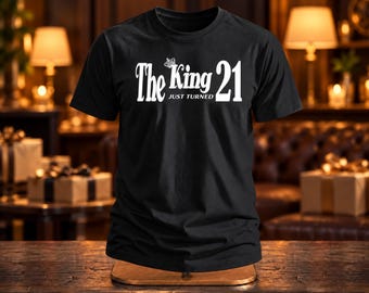 May include: A black short-sleeved t-shirt featuring white text that reads The King JUST TURNED 21 with a small crown icon above the word King. The garment has a crew neckline and a standard fit, displayed on a flat surface.