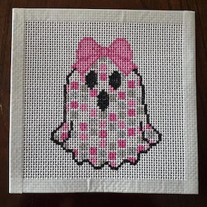 May include: A square needlepoint canvas featuring a ghost design stitched with a pink and gray checkered pattern. The ghost has two black eyes and a black mouth, and it is wearing a large pink bow on its head. The canvas has white taped edges.