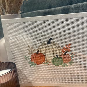 May include: A rectangular needlepoint canvas featuring a cross-stitched design of four pumpkins in varying sizes and colors, including orange, cream, and green, accented with small leaves and stems, set against a white grid background.