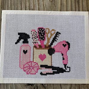 May include: A needlepoint canvas featuring a cross-stitch design of hair salon tools. The pattern includes a pink spray bottle with a heart, a hairbrush, scissors, a comb, a pink hair dryer, and a flat iron, all arranged around a central container.
