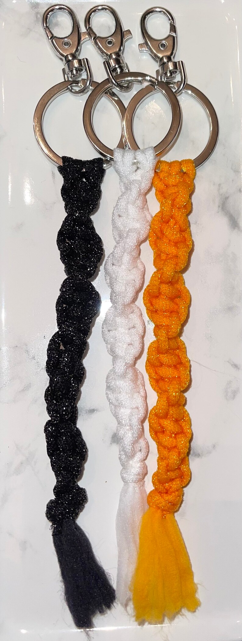 May include: Three keychains with black, white and yellow braided tassels. Each keychain has a silver metal ring and a silver metal clasp.