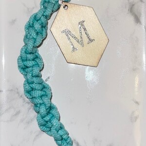 May include: A teal blue braided keychain with a silver key ring and a hexagonal wooden tag with a silver glitter letter "M" on it.