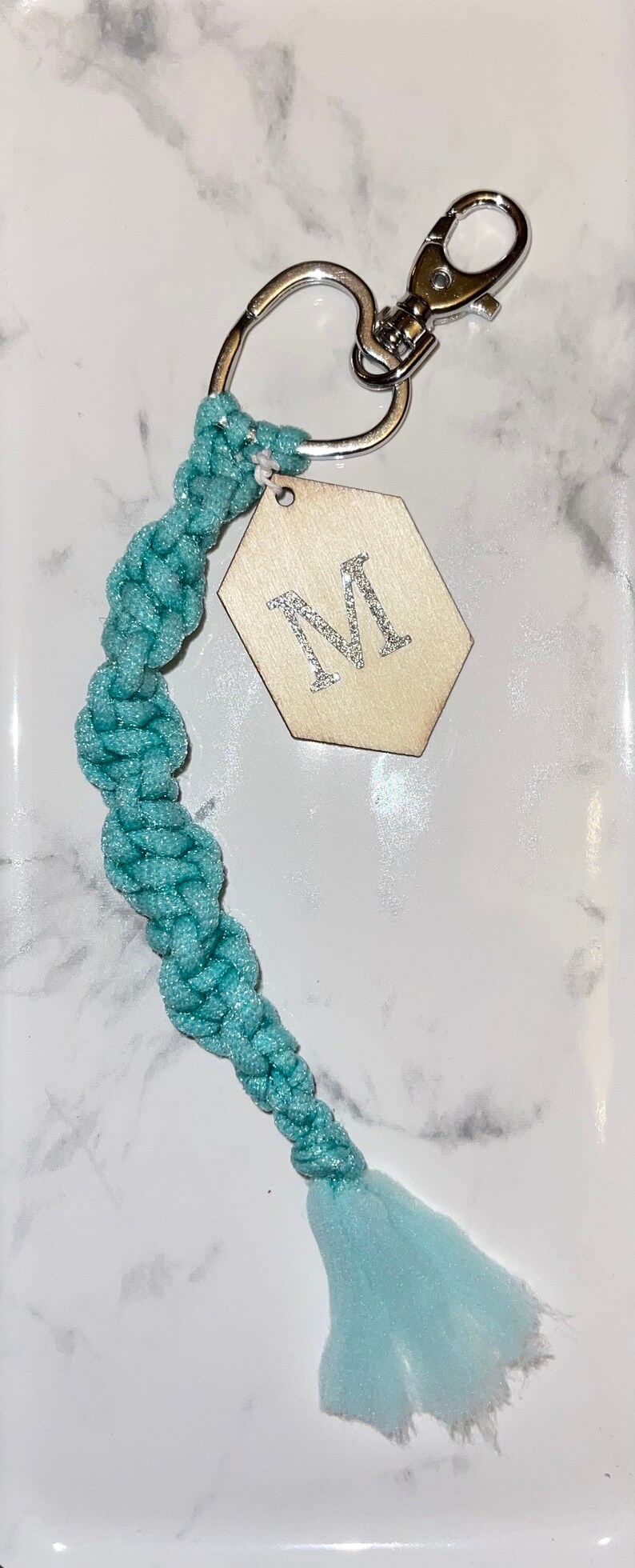 May include: A teal blue braided keychain with a silver key ring and a hexagonal wooden tag with a silver glitter letter "M" on it.