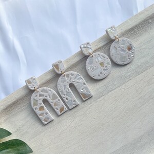 May include: Three pairs of white mosaic-style earrings with gold-colored hooks are displayed on a light wooden tray. The earrings have different shapes: arch, circle, and square. The mosaic design features iridescent pieces.