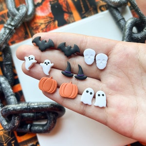 Op de afbeelding: Een set Halloween-thema polymer clay oorbellen. De oorbellen hebben verschillende ontwerpen, waaronder vleermuizen, geesten, pompoenen en schedels. De oorbellen zijn allemaal wit, zwart en oranje.