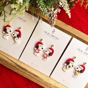 May include: Three pairs of handmade polymer clay earrings featuring a cartoon dog wearing a Santa hat and holding a gift. The earrings are displayed on white cards with the text "Lilac and Lili Handmade with Love" and the social media handle "@lilacandili".