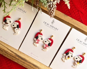 Puede incluir: Tres pares de pendientes hechos a mano de arcilla polimérica con un perro de dibujos animados que lleva un gorro de Papá Noel y sostiene un regalo. Los pendientes se muestran en tarjetas blancas con el texto "Lilac and Lili Handmade with Love" y el nombre de usuario de las redes sociales "@lilacandili".