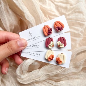 May include: Three pairs of polymer clay earrings featuring miniature pumpkins with flowers. The pumpkins are in shades of orange, cream, and burgundy. The flowers are in shades of cream, burgundy, and orange. The earrings are handmade with love.