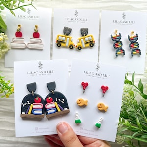 Może przedstawiać: Pięć kartek z tekstem "Lilac and Lili Handmade with Love" i różnymi kolczykami z polimerowej gliny w szkolnym stylu. Kolczyki obejmują stos książek z czerwonym jabłkiem na górze, żółty autobus szkolny, łańcuch DNA, czarną tablicę z książkami i jabłkiem, czerwone nożyczki, złotą rybkę i zieloną probówkę.