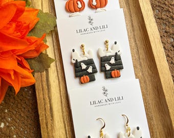 Könnte beinhalten: Halloween-Ohrringe mit Snoopy-Designs und Kürbissen. Die Ohrringe sind aus Polymer Clay gefertigt und haben goldfarbene Haken. Die Designs umfassen Snoopy auf Kürbissen, Snoopy in einem Hundehütte mit Geistern und Snoopy auf Kürbislaternen. Die Karten lesen "LILAC AND LILI HANDMADE WITH LOVE."