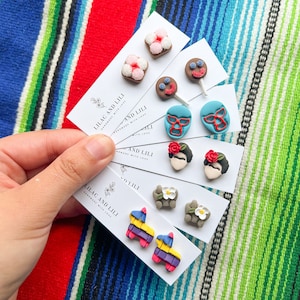 Puede incluir: Un conjunto de pendientes de arcilla polimérica de colores en tarjetas blancas con el texto "Lilac and Lili Handmade with Love". Los pendientes presentan diseños como una piñata, una máscara de luchador, una flor, una calavera de azúcar y una paleta.