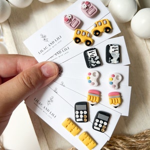 May include: Set of six pairs of colorful clay earrings with school-themed designs, including a backpack, school bus, notebook, paint palette, pencil, and calculator. The earrings are displayed on white cards with the text "Lilac and Lili Handmade with Love".