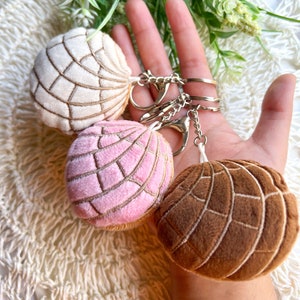 May include: Three plush keychains shaped like Mexican sweet bread, known as conchas. The conchas are white, pink, and brown. The keychains are attached to a silver chain.