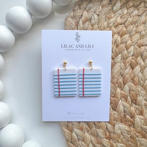 May include: A pair of blue and white notepad-shaped earrings with red lines and gold hooks. The earrings are on a white card with the text "Lilac and Lili Handmade with Love" and the social media handle "@lilacandili" and website "www.lilacandili.etsy.com".