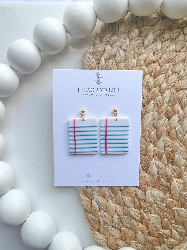 May include: A pair of blue and white notepad-shaped earrings with red lines and gold hooks. The earrings are on a white card with the text "Lilac and Lili Handmade with Love" and the social media handle "@lilacandili" and website "www.lilacandili.etsy.com".