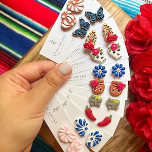 Könnte beinhalten: Ein Set farbenfroher Ohrringe aus Polymer Clay mit verschiedenen Designs, darunter Herzen, Blumen, Schädel, Schmetterlinge und Chilischoten. Die Ohrringe sind handgefertigt und werden auf weißen Karten mit dem Text "Lilac and Lili" und "Handmade with Love" geliefert.